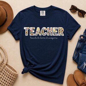May include: Navy blue t-shirt featuring the word "TEACHER" in white block letters, filled with colourful floral designs. The text "teach love inspire" is below. The shirt is folded at the bottom.