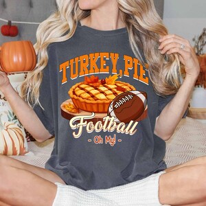 Op de afbeelding: Donkergrijs T-shirt met de tekst "TURKEY, PIE, Football - Oh My!" in oranje en wit. De afbeelding bevat een taart, een voetbal en herfstbladeren. Het shirt heeft een casual stijl.