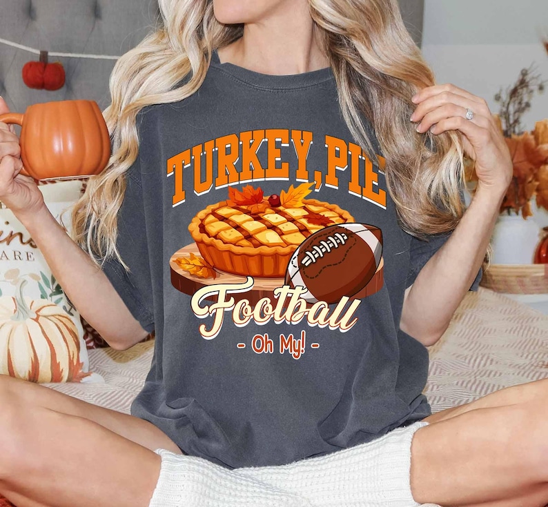 Op de afbeelding: Donkergrijs T-shirt met de tekst "TURKEY, PIE, Football - Oh My!" in oranje en wit. De afbeelding bevat een taart, een voetbal en herfstbladeren. Het shirt heeft een casual stijl.