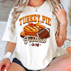 Op de afbeelding: Wit T-shirt met een afbeelding van een taart, een voetbal en de woorden "TURKEY, PIE Football - Oh My!". Het ontwerp bevat oranje en bruine kleuren, met herfstbladeren. Een leuk, feestelijk ontwerp voor het herfstseizoen.