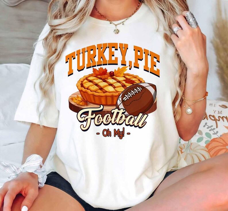 Op de afbeelding: Wit T-shirt met een afbeelding van een taart, een voetbal en de woorden "TURKEY, PIE Football - Oh My!". Het ontwerp bevat oranje en bruine kleuren, met herfstbladeren. Een leuk, feestelijk ontwerp voor het herfstseizoen.