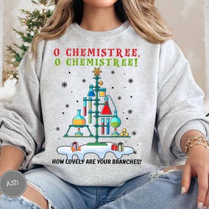 May include: A light gray sweatshirt with a Christmas tree design made of laboratory equipment. The text "O Chemistree, O Chemistree! How lovely are your branches!" is printed in red and green. Snowflakes and gifts are also featured.