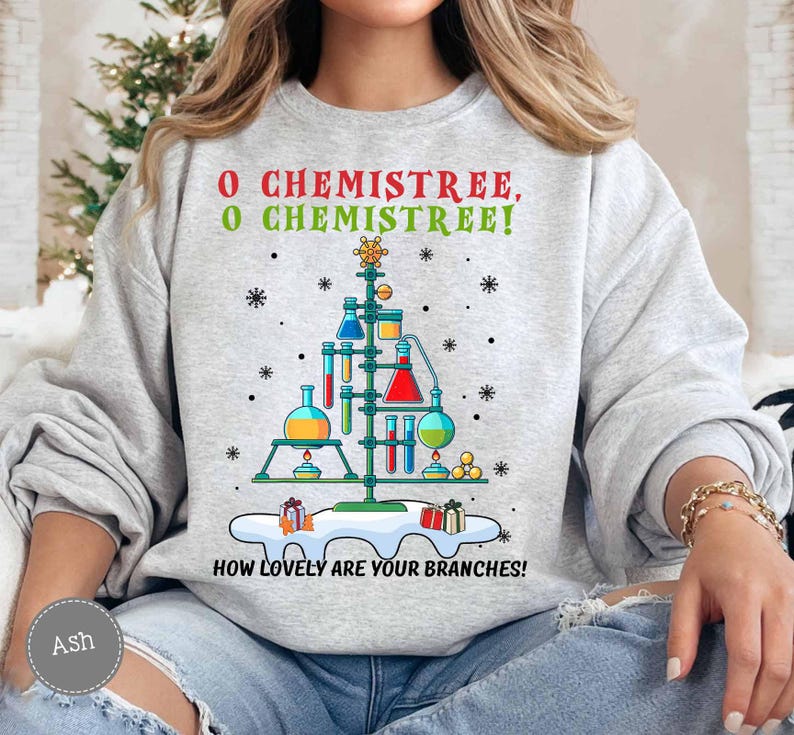 May include: A light gray sweatshirt with a Christmas tree design made of laboratory equipment. The text "O Chemistree, O Chemistree! How lovely are your branches!" is printed in red and green. Snowflakes and gifts are also featured.