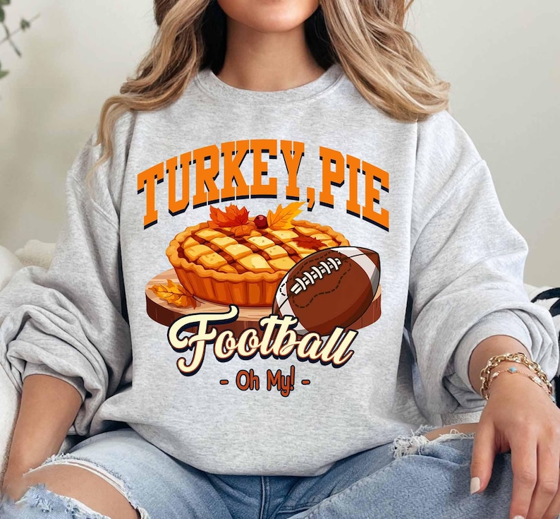 Op de afbeelding: Een lichtgrijs sweatshirt met een grafisch ontwerp. Het ontwerp bevat de woorden "TURKEY, PIE Football - Oh My!" boven een illustratie van een taart en een voetbal. De taart staat op een houten plank met herfstbladeren.