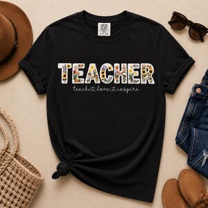 May include: Black t-shirt featuring the word "TEACHER" in white, floral-filled letters. The text "teach, love, inspire" is written below in a cursive font. The shirt is styled with sunglasses, a hat, and jeans.