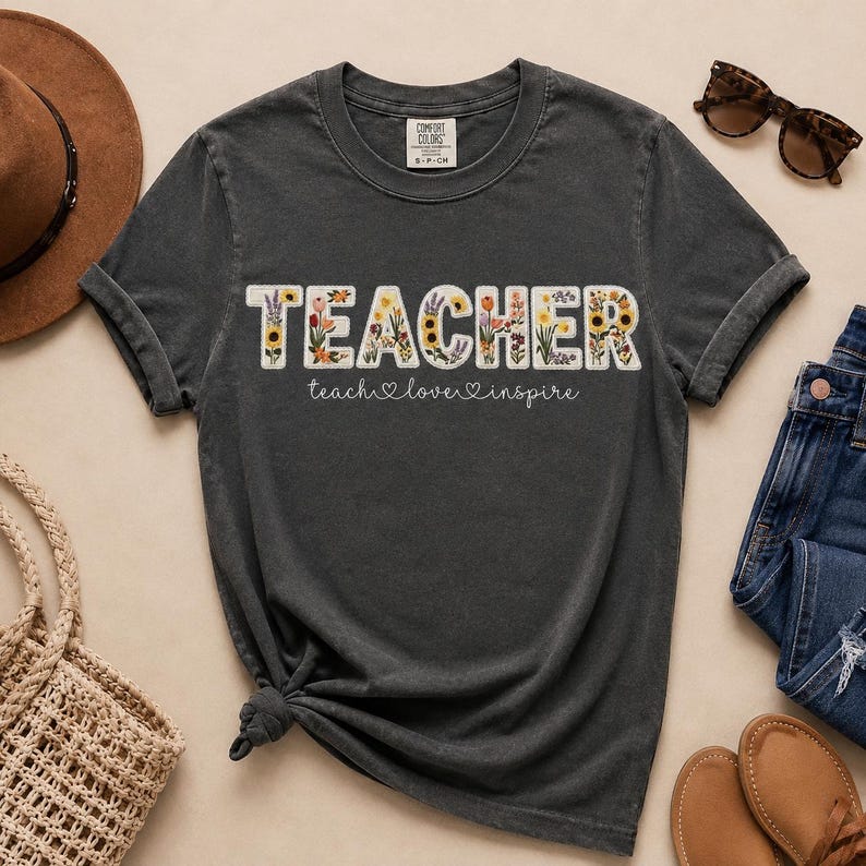 May include: Dark grey t-shirt featuring the word "TEACHER" in a floral design. The text "teach, love, inspire" is below the word "TEACHER". The shirt is a Comfort Colors brand, and is paired with jeans, sunglasses, and a hat.