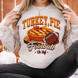 Op de afbeelding: Een cr&egrave;mekleurige sweatshirt met de tekst "TURKEY, PIE Football - Oh My!" in oranje en zwart. De afbeelding bevat een taart, een voetbal en herfstbladeren. De sweatshirt is een casual, seizoensgebonden ontwerp.