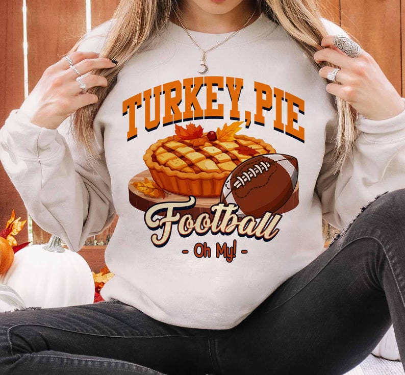 Op de afbeelding: Een cr&egrave;mekleurige sweatshirt met de tekst "TURKEY, PIE Football - Oh My!" in oranje en zwart. De afbeelding bevat een taart, een voetbal en herfstbladeren. De sweatshirt is een casual, seizoensgebonden ontwerp.