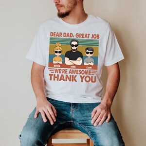 May include: White t-shirt with a vintage-style graphic that says "Dear Dad, Great Job" and features a cartoon dad and two kids. The text below the graphic says "We're Awesome Thank You" and includes the names "Olivia, John, Liam".