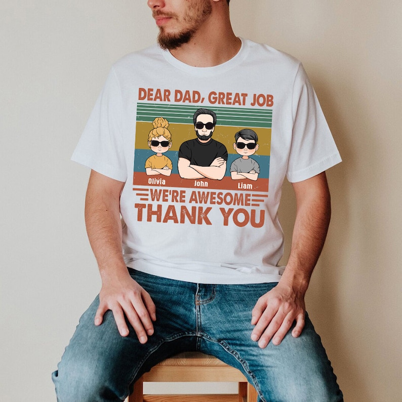 May include: White t-shirt with a vintage-style graphic that says "Dear Dad, Great Job" and features a cartoon dad and two kids. The text below the graphic says "We're Awesome Thank You" and includes the names "Olivia, John, Liam".