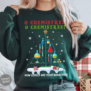 May include: Forest green sweatshirt with a Christmas tree design made of laboratory equipment. The text "O Chemistree! How lovely are your branches!" is printed on the front, along with snowflakes and gift boxes.