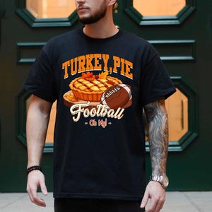 Op de afbeelding: Zwart t-shirt met een afbeelding van een kalkoentaart en een voetbal. De tekst op het shirt luidt "TURKEY, PIE Football - Oh My!" in oranje en wit. Het ontwerp bevat herfstbladeren.