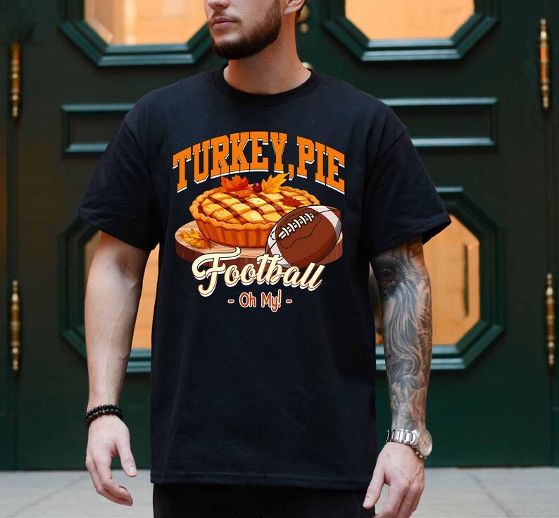 Op de afbeelding: Zwart t-shirt met een afbeelding van een kalkoentaart en een voetbal. De tekst op het shirt luidt "TURKEY, PIE Football - Oh My!" in oranje en wit. Het ontwerp bevat herfstbladeren.