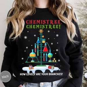 May include: Black sweatshirt with a Christmas tree design made of laboratory glassware. The text "CHEMISTREE, CHEMISTREE! HOW LOVELY ARE YOUR BRANCHES!" is printed in green and white. Snowflakes and gift boxes complete the festive look.
