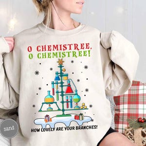 May include: A sand-colored sweatshirt with a Christmas tree design made of laboratory equipment. The text "O CHEMISTREE, O CHEMISTREE! HOW LOVELY ARE YOUR BRANCHES!" is printed above and below the tree. Snowflakes and gifts are also included.