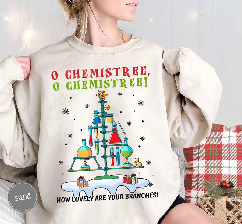 May include: A sand-colored sweatshirt with a Christmas tree design made of laboratory equipment. The text "O CHEMISTREE, O CHEMISTREE! HOW LOVELY ARE YOUR BRANCHES!" is printed above and below the tree. Snowflakes and gifts are also included.
