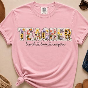 May include: A pale pink t-shirt featuring the word "TEACHER" in a floral design. The words "teach love inspire" are written below in cursive. The shirt has a crew neck and short sleeves.