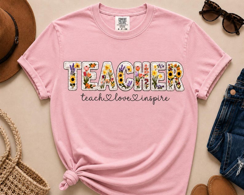 May include: A pale pink t-shirt featuring the word "TEACHER" in a floral design. The words "teach love inspire" are written below in cursive. The shirt has a crew neck and short sleeves.