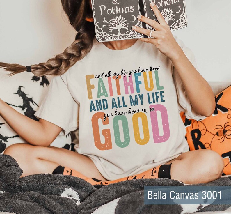 May include: A white t-shirt with a colorful graphic that reads "and all my life you have been FAITHFUL AND ALL MY LIFE you have been so, so GOOD". The shirt is on a person sitting on a couch with a black and orange pillow.