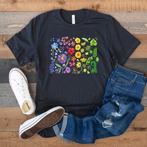 May include: A black t-shirt with a rainbow floral print. The print features a variety of flowers and leaves in different shades of red, orange, yellow, green, blue, and purple. The shirt is paired with blue jeans and white sneakers.