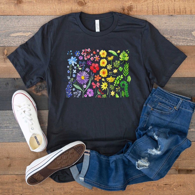 May include: A black t-shirt with a rainbow floral print. The print features a variety of flowers and leaves in different shades of red, orange, yellow, green, blue, and purple. The shirt is paired with blue jeans and white sneakers.