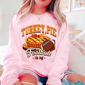 Op de afbeelding: Roze sweatshirt met de woorden "TURKEY, PIE" in oranje letters boven een afbeelding van een taart en een voetbal. De afbeelding toont een taart op een houten plank en een bruine voetbal. De woorden "Football - Oh My!" staan eronder.