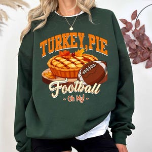 Op de afbeelding: Donkergroene sweatshirt met een afbeelding van een taart, een voetbal en de tekst "TURKEY, PIE Football - Oh My!". De taart is goudbruin met herfstbladeren. De voetbal is bruin en wit.