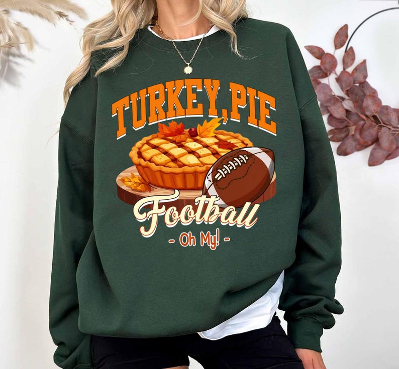 Op de afbeelding: Donkergroene sweatshirt met een afbeelding van een taart, een voetbal en de tekst "TURKEY, PIE Football - Oh My!". De taart is goudbruin met herfstbladeren. De voetbal is bruin en wit.