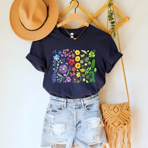 May include: A navy blue t-shirt with a rainbow floral print. The shirt is hanging on a wooden hanger with a brown hat and a woven bag.