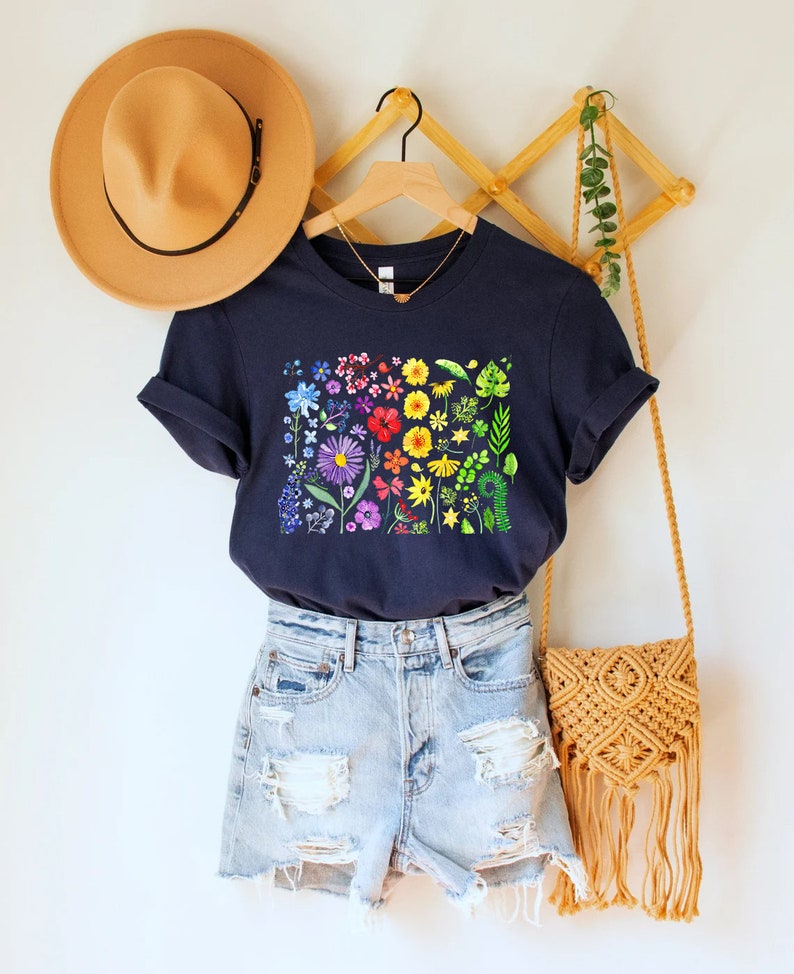 May include: A navy blue t-shirt with a rainbow floral print. The shirt is hanging on a wooden hanger with a brown hat and a woven bag.