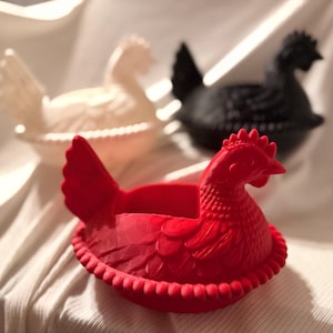 May include: Three ceramic hen-shaped containers in red, white, and black. The red hen is in the foreground, with a detailed comb and feather design. The white and black hens are in the background, with a similar design. These decorative containers are ideal for kitchen decor.
