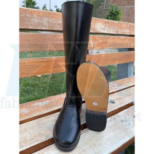 May include: A pair of black leather riding boots. One boot stands upright, while the other is positioned to show the sole. The boots are on a wooden bench, with a green background.