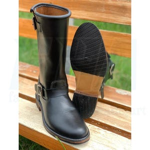 Puede incluir: Botas de moto de cuero negro con detalles de hebillas. Una bota está en posición vertical, la otra se muestra con la suela hacia arriba. Las botas tienen punta redonda y tacón bajo. Las suelas son de goma oscura.