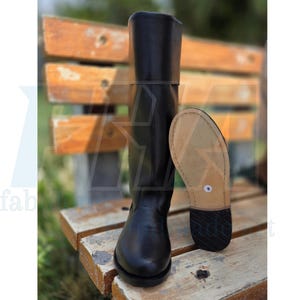 May include: A tall black leather boot with a rounded toe and a wide cuff at the top. Beside the boot stands a second boot, showing its light tan leather sole with a small white circular size label and a black heel with a textured tread.