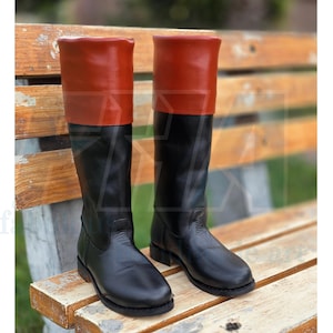 May include: A pair of black leather riding boots with a red leather cuff at the top. The boots are knee-high and have a classic design. They are displayed on a wooden bench.