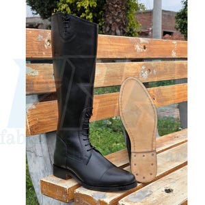 Puede incluir: Bota de montar de cuero negro con cordones en la parte delantera y puntera redondeada. La bota es alta, llega hasta la rodilla. La suela es de color marrón claro con costuras visibles y tachuelas metálicas. La bota se muestra junto a su suela.