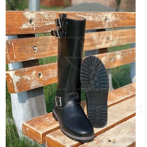 May include: Black leather knee-high boots with multiple silver buckles and a lug sole. One boot is upright, and the other is lying on its side, showcasing the tread. The boots are on a wooden bench.