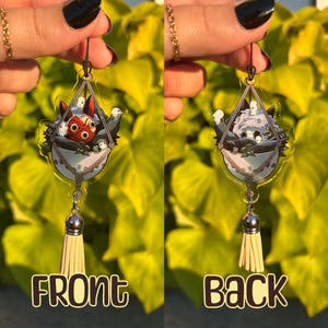 May include: Clear acrylic earrings featuring a cartoon design. The earrings have a triangular shape with a macrame-style basket design. The front earring features a red and white character, while the back earring shows a white and gray character. Each earring has a silver-colored charm and a beige tassel.
