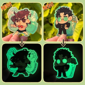 May include: Two glow-in-the-dark acrylic charms featuring anime-style characters. One shows a person with brown hair and a red bow, the other a person with dark hair and round glasses. Both designs glow bright green in the dark and include the text NEW.
