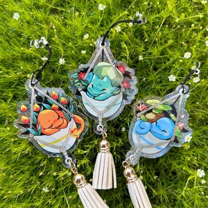 Op de afbeelding: Drie heldere acryl bedels met stripfiguren in hangende plantenbakken en witte kwastjes. De figuren zijn oranje, blauw en groen, omringd door bloemen. Elke bedel heeft een zwart koord om op te hangen.