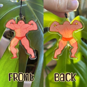 May include: A clear acrylic charm featuring a muscular figure in a red swimsuit. The front of the charm has the word "FRONT" in gold lettering, while the back shows the figure's back with the word "BACK". The charm is suspended by a black string.