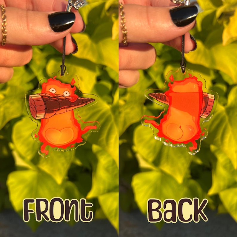 May include: A clear acrylic charm featuring a cartoon character in shades of orange and red, with a brown object. The charm has a black string for hanging. The text "FRONT" and "BACK" are visible.