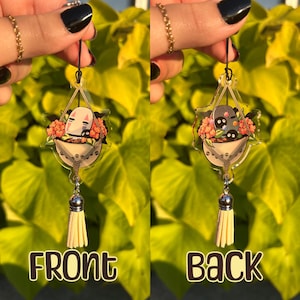 May include: A clear acrylic charm featuring a cartoon character in a basket of flowers, with a cream-colored tassel. The front of the charm says "FRONT" and the back says "BACK". The charm is suspended by a black string.