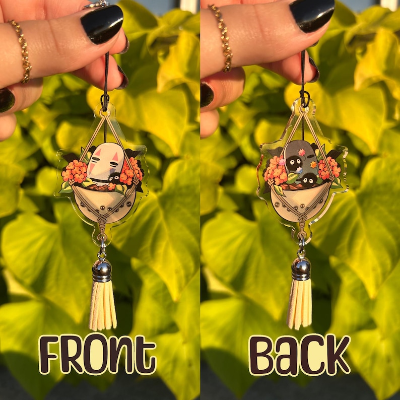 May include: A clear acrylic charm featuring a cartoon character in a basket of flowers, with a cream-colored tassel. The front of the charm says "FRONT" and the back says "BACK". The charm is suspended by a black string.