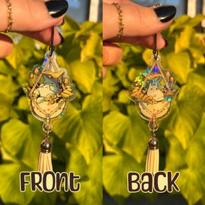 May include: A clear acrylic charm featuring a cartoon character in a hammock with mushrooms and leaves. The charm has a silver-colored tassel and the word "FRONT" is printed on the front. The back of the charm has the word "BACK".