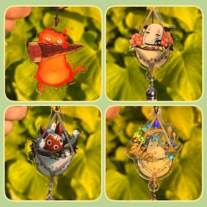 May include: Four clear acrylic charms featuring characters from a popular animated film. The charms include a fiery creature, a masked spirit, a forest spirit, and a gray creature, each suspended from a black cord. The charms are set against a green leafy background.