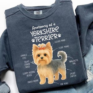 以下が含まれることがあります： 「Anatomy of a Yorkshire Terrier」の文字とヨークシャーテリアのイラストがプリントされたネイビーブルーのスウェットシャツ。犬の解剖学的構造がユーモラスにラベル付けされています。スウェットシャツは、青いジーンズと組み合わせて表示されています。