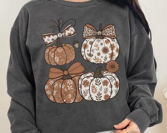 May include: Dark gray sweatshirt featuring a fall-themed design. The graphic includes four pumpkins with floral and bow details in shades of brown, cream, and tan. The sweatshirt has a crew neck and long sleeves, perfect for autumn.