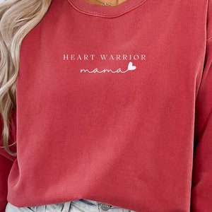 May include: Crimson-colored sweatshirt with the text "HEART WARRIOR mama" in white script. The sweatshirt has a crew neck and long sleeves. The model is wearing light blue denim shorts. The word "CRIMSON" is in a circle on the bottom right.