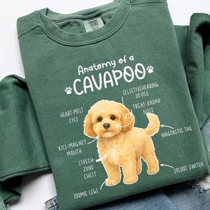 Op de afbeelding: Een donkergroene sweatshirt met de tekst "Anatomy of a Cavapoo" en een diagram van een Cavapoo-hond. Het diagram labelt de kenmerken van de hond, waaronder "heart-melt eyes" en "wagtail tail". De sweatshirt ligt op een spijkerbroek.
